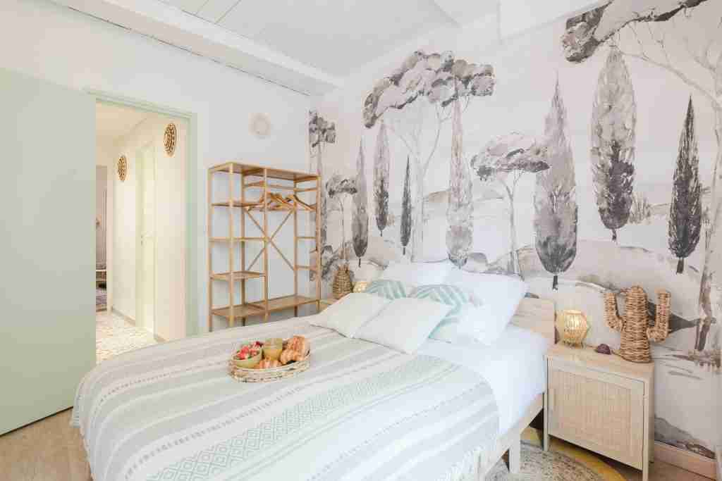 Chambre lumineuse et moderne avec lit double décoré de coussins verts et blancs. Papier peint nature avec arbres. Étagère en bois et table de chevet complètent le décor douillet.