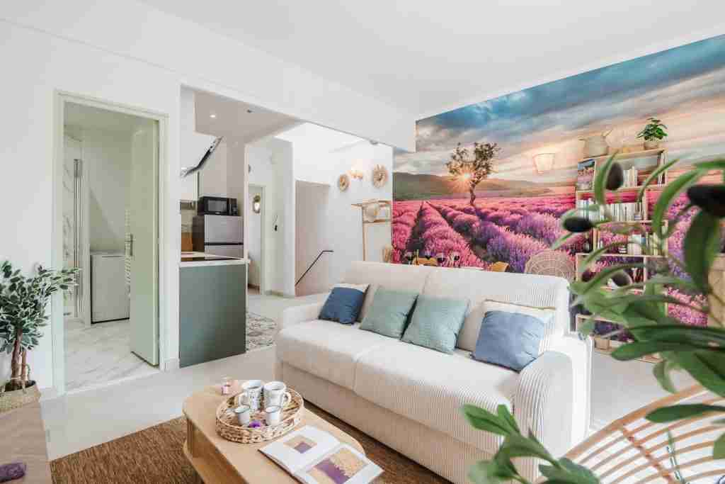 Salon lumineux avec canapé blanc et coussins colorés devant un mur avec une fresque de lavande, offrant une ambiance relaxante dans une maison moderne et accueillante.