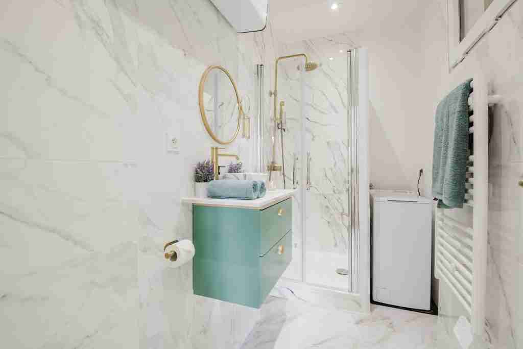 Salle de bain moderne avec douche en verre et carrelage en marbre élégant. Meuble vasque vert avec miroir rond et accessoires dorés pour une esthétique raffinée.