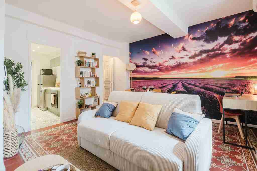 Salon moderne avec mur en papier peint représentant un coucher de soleil sur un champ de lavande canapé blanc coussins colorés étagères et bureau aménagés éclairage doux.
